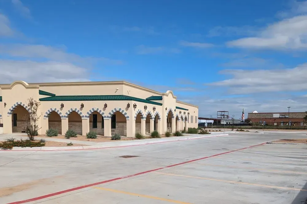 Abandoned beige building with arches beside an empty concrete lot under a blue sky