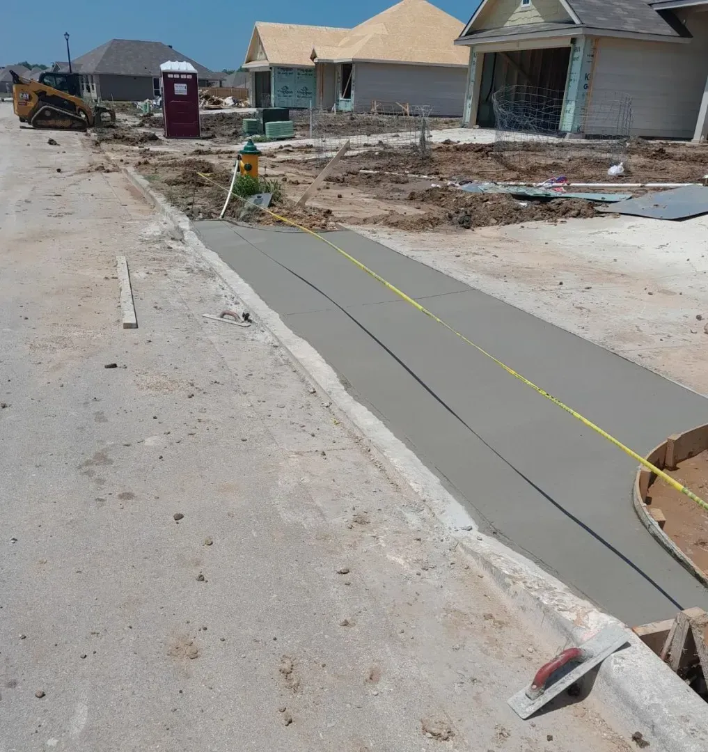Damaged sidewalk at a suburban street under construction, with debris and unfinished concrete near houses.