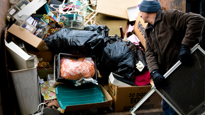 A man is pushing a screen door into a dumpster filled with garbage.