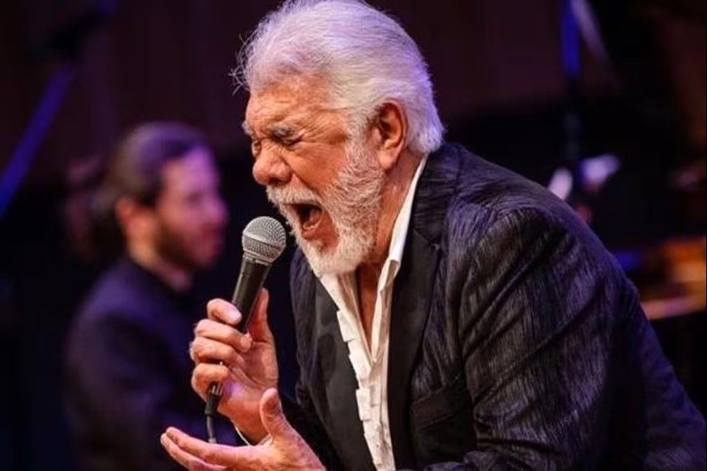 Hombre con cabello y barba blancos cantando en un micrófono en el escenario, con un miembro de la banda en el fondo.