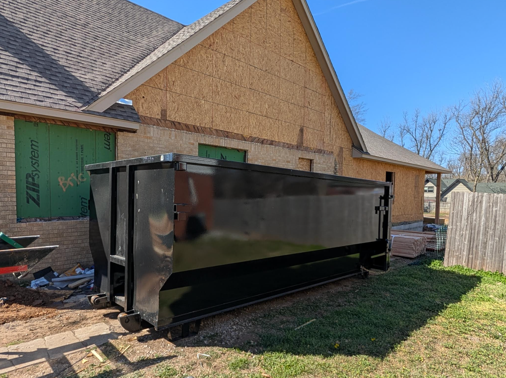Two large, gray trash containers with closed lids on a paved surface, with trees in the background.