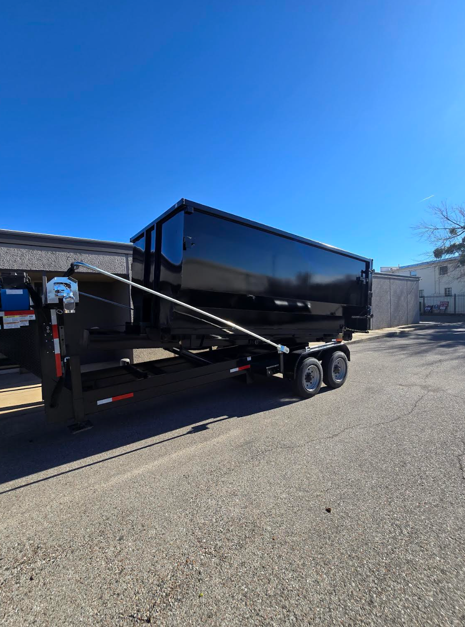 Green dumpster filled with debris sits in a driveway next to a house with a green lawn and trees.