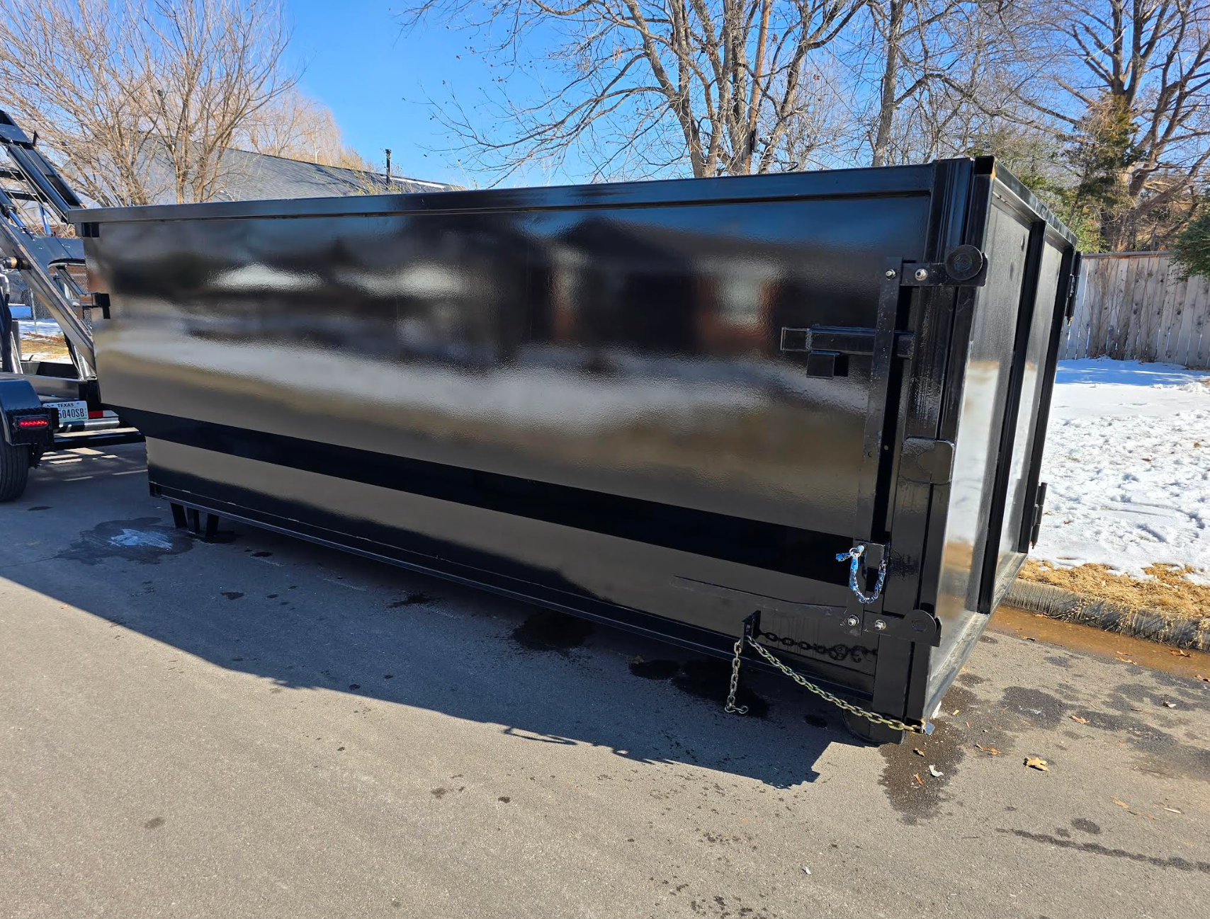 Four dark dumpsters lined up on a street, with houses and trees in the background under a cloudy sky.