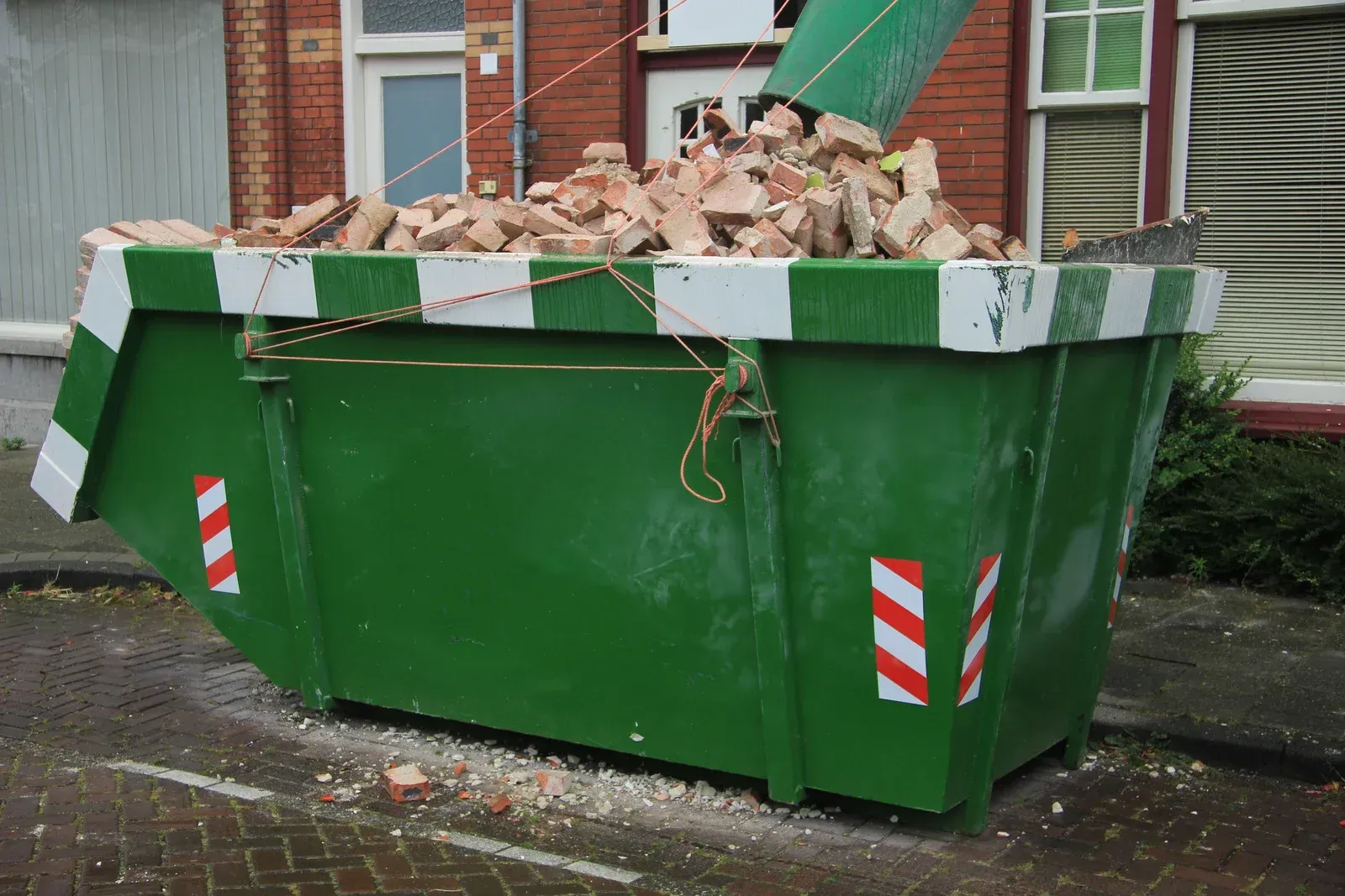 Green dumpster overflowing with bricks, on a brick road in front of a house.