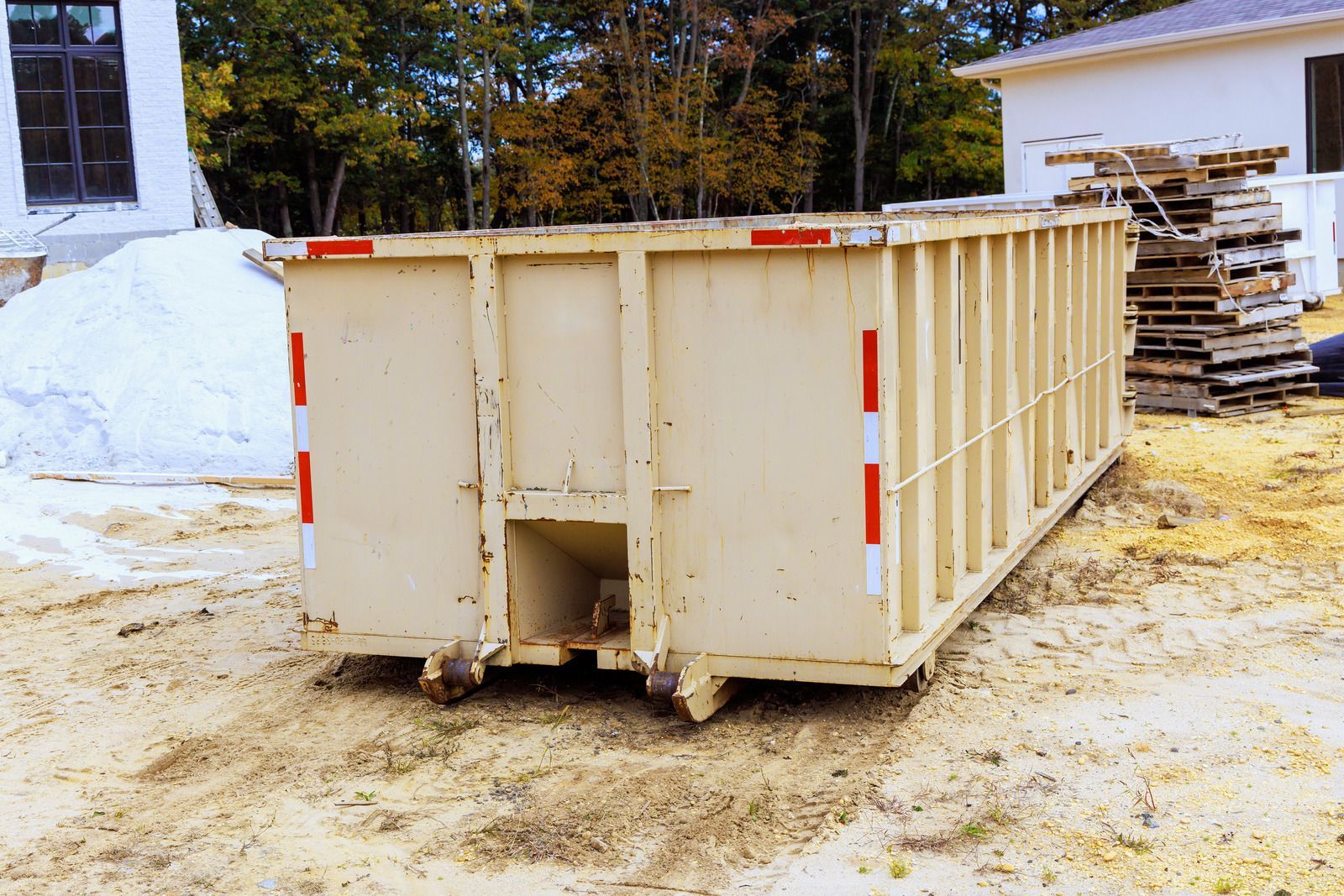 Beige construction dumpster on a sandy construction site, with a building under construction in the background.