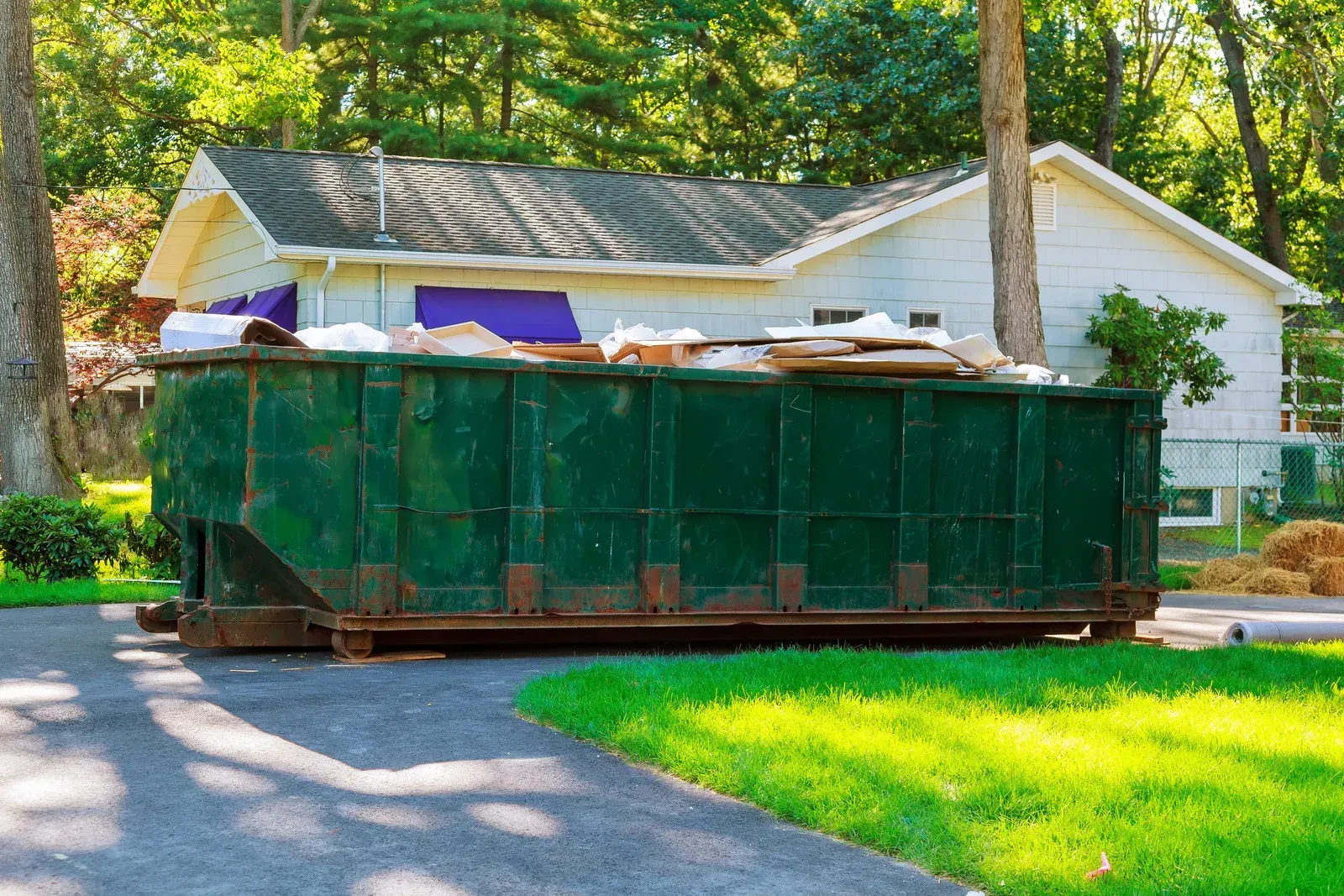 Green dumpster filled with debris sits in a driveway next to a house with a green lawn and trees.