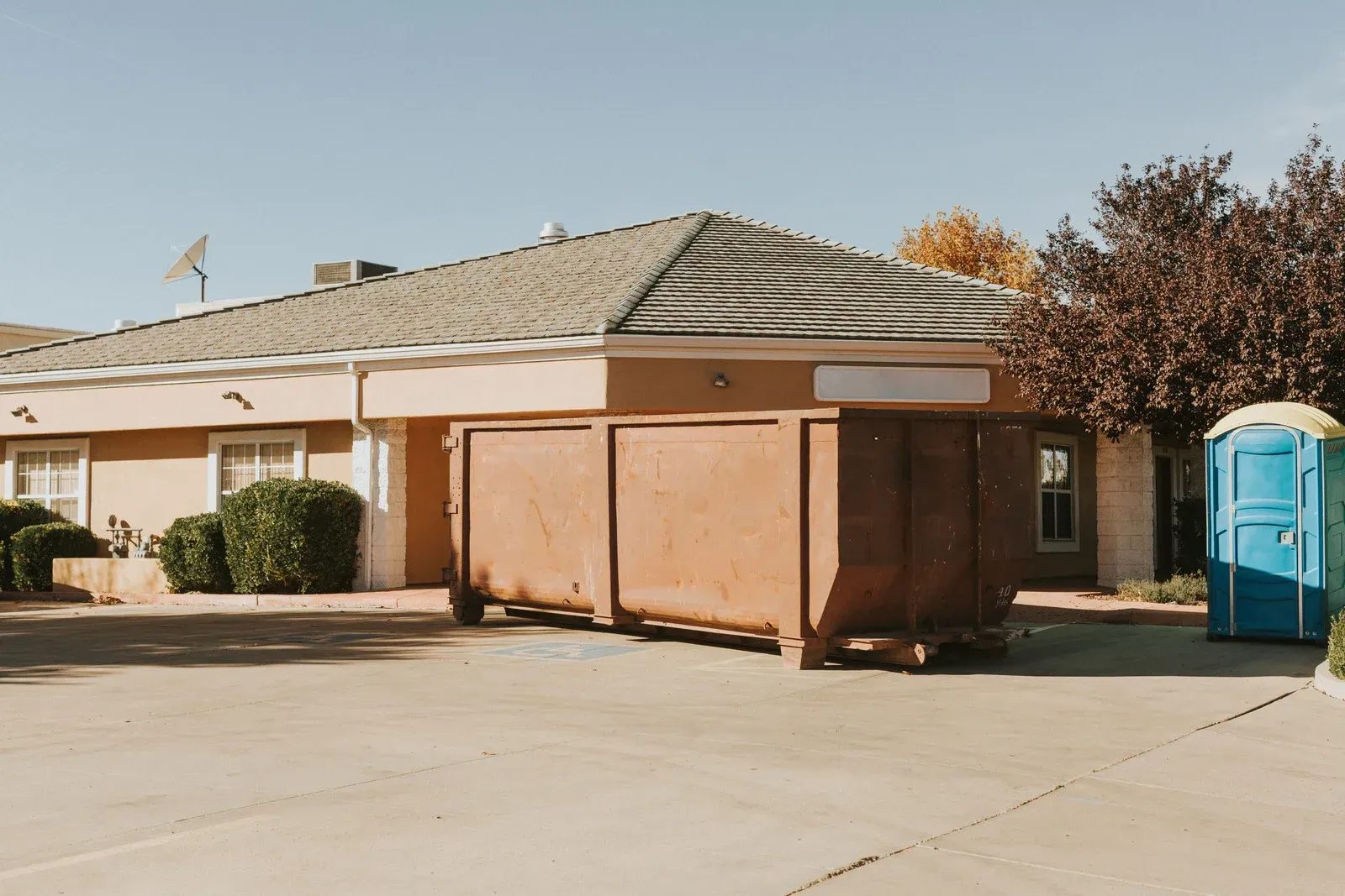 Tan building with brown dumpster and blue portable toilet in a parking lot.