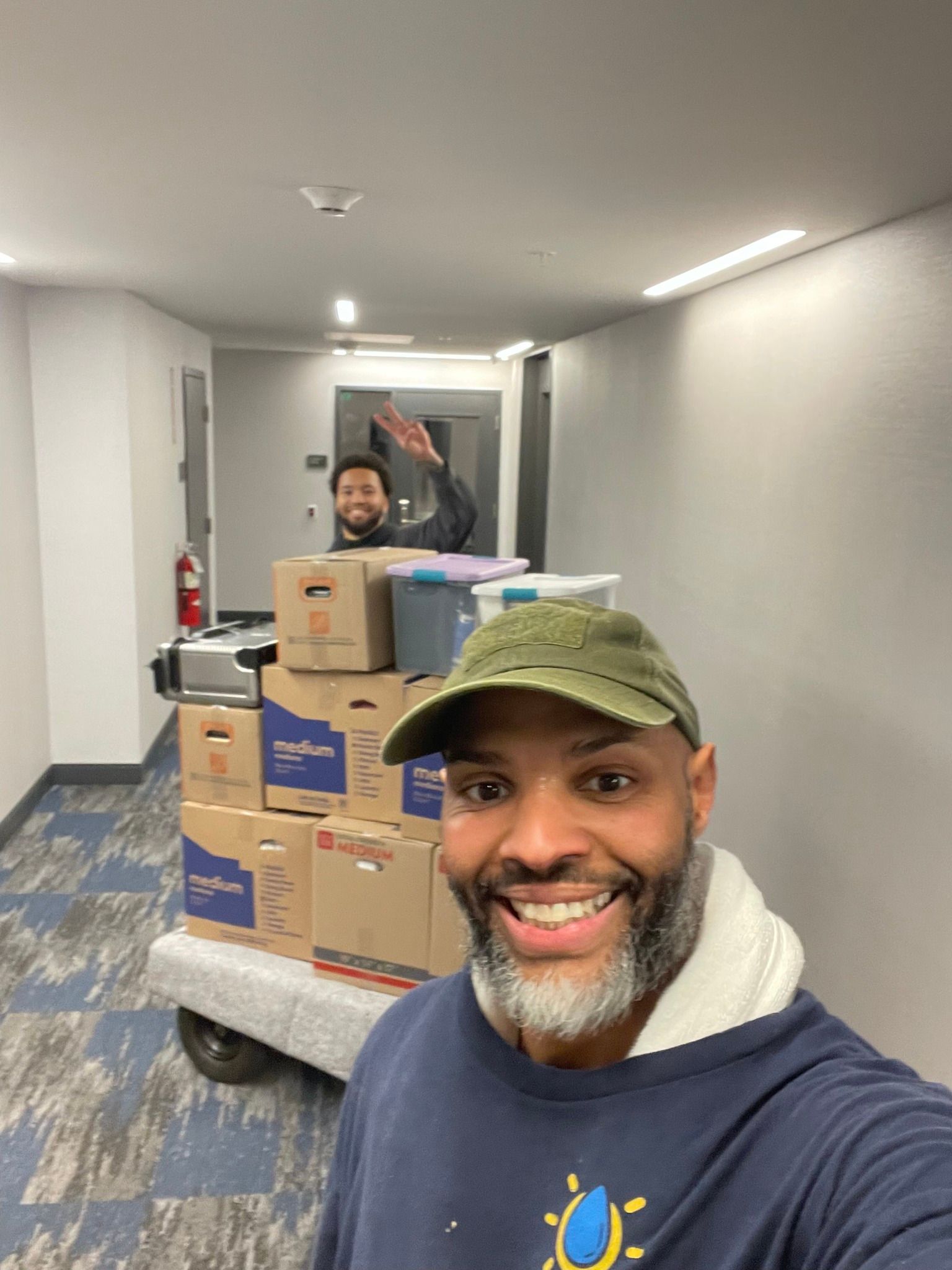 Man smiling, taking a selfie with boxes on a cart, another man waving from behind in a hallway.