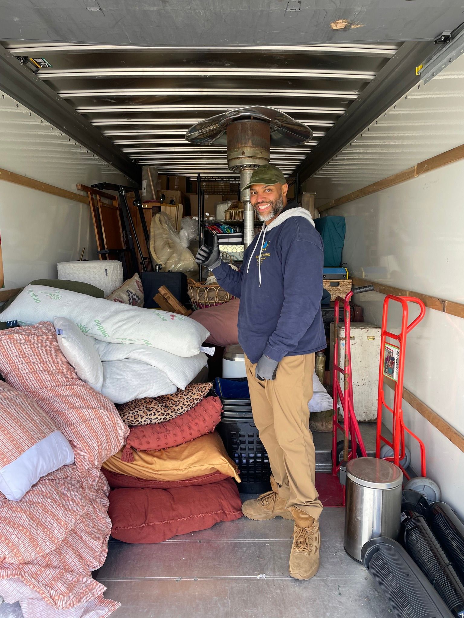 Man stands in a packed moving truck, smiling.  Pillows, furniture, and a heater are visible.