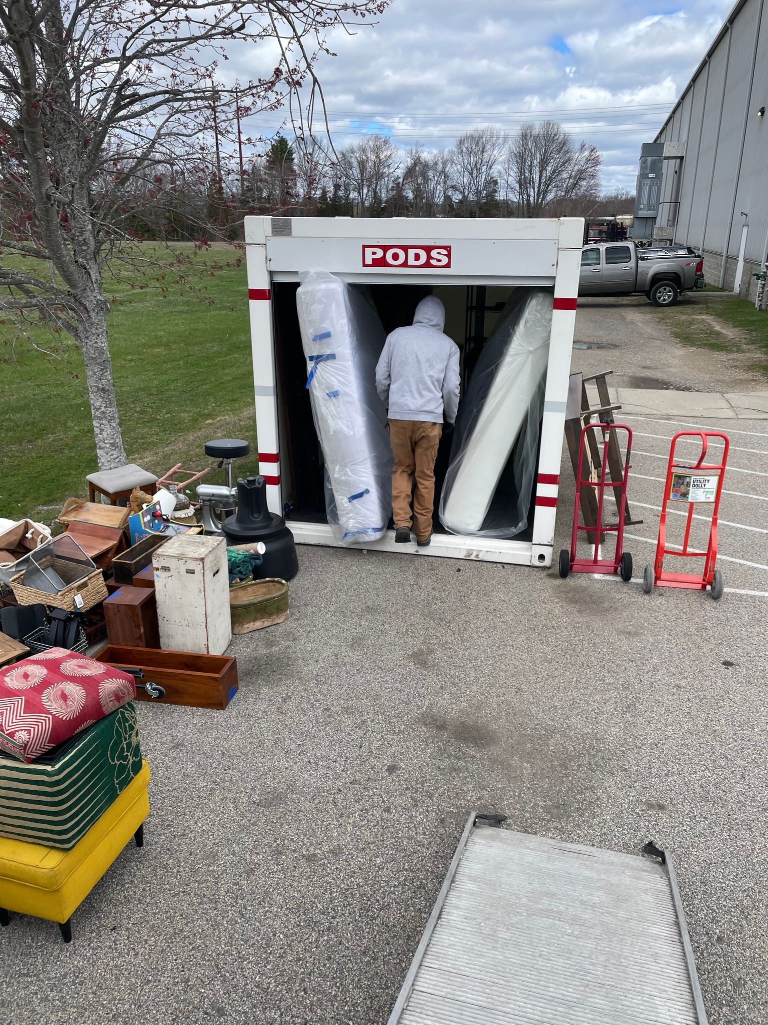 A person loads a PODS storage container with mattresses. Outdoor setting with other furniture nearby.