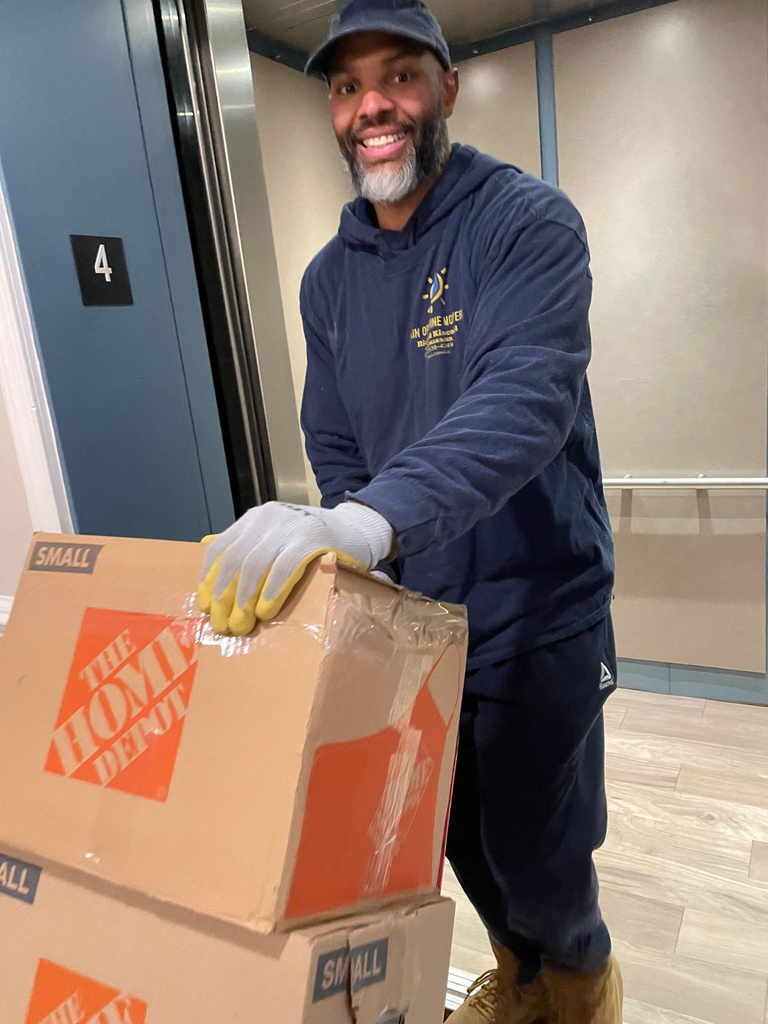 Man in blue hoodie and cap, wearing gloves, carrying two Home Depot boxes in elevator.