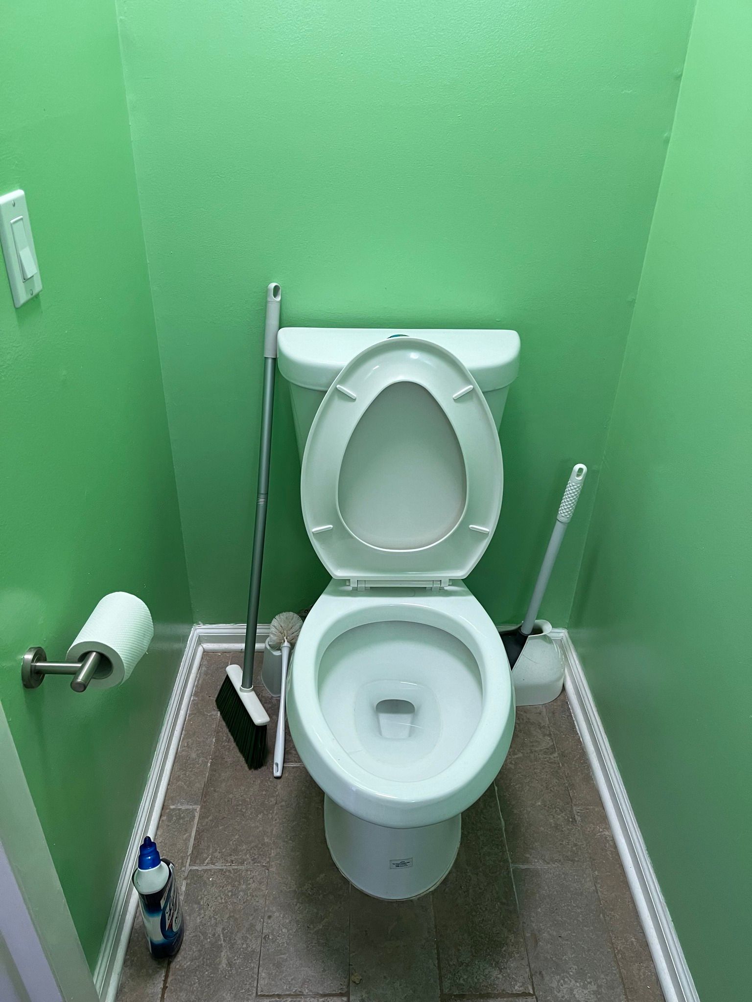 Toilet in a small green-walled bathroom with toilet paper, cleaning brush, and broom on the floor.