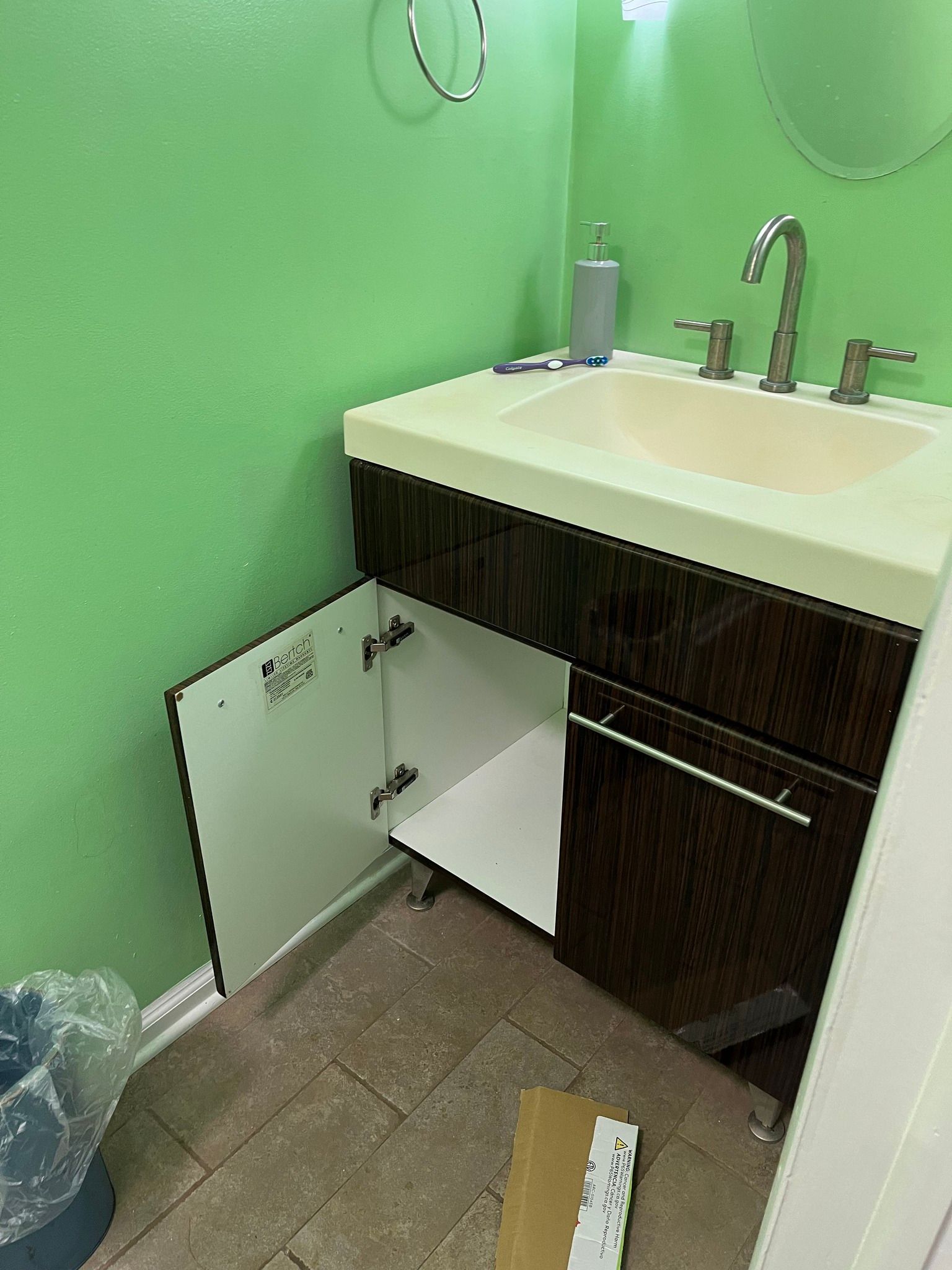 Bathroom vanity with open cabinet door, brown cabinet, beige sink, green wall, tiled floor.