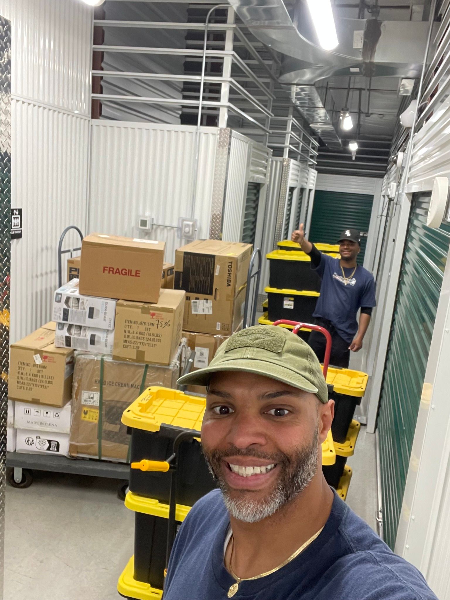Two men in storage unit with boxes, smiling, near a cart stacked with yellow bins.