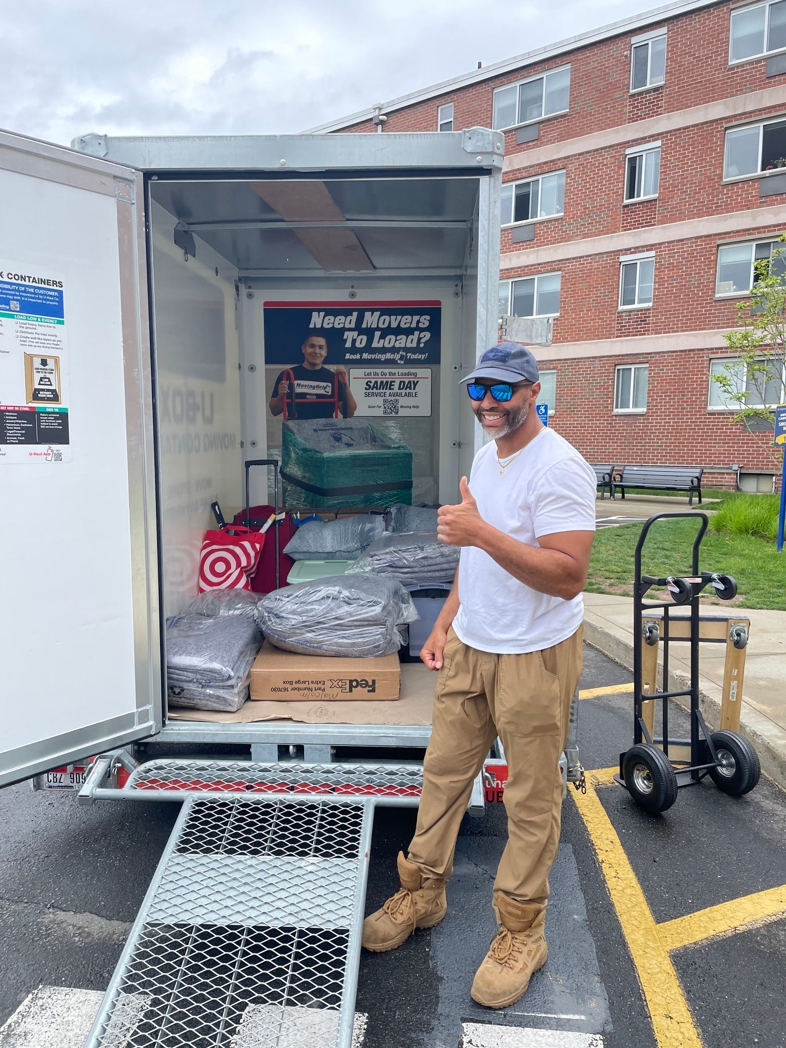 Man by an open moving truck, giving a thumbs-up. Truck holds donations. Apartment building in the background.