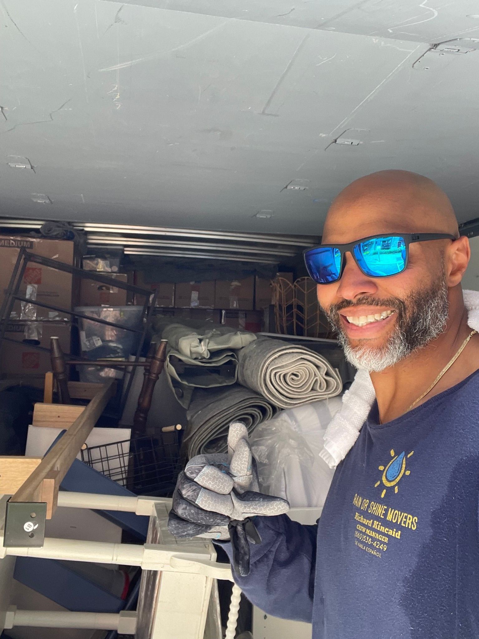 Smiling man in blue sunglasses in a cluttered cargo space, giving a thumbs up.