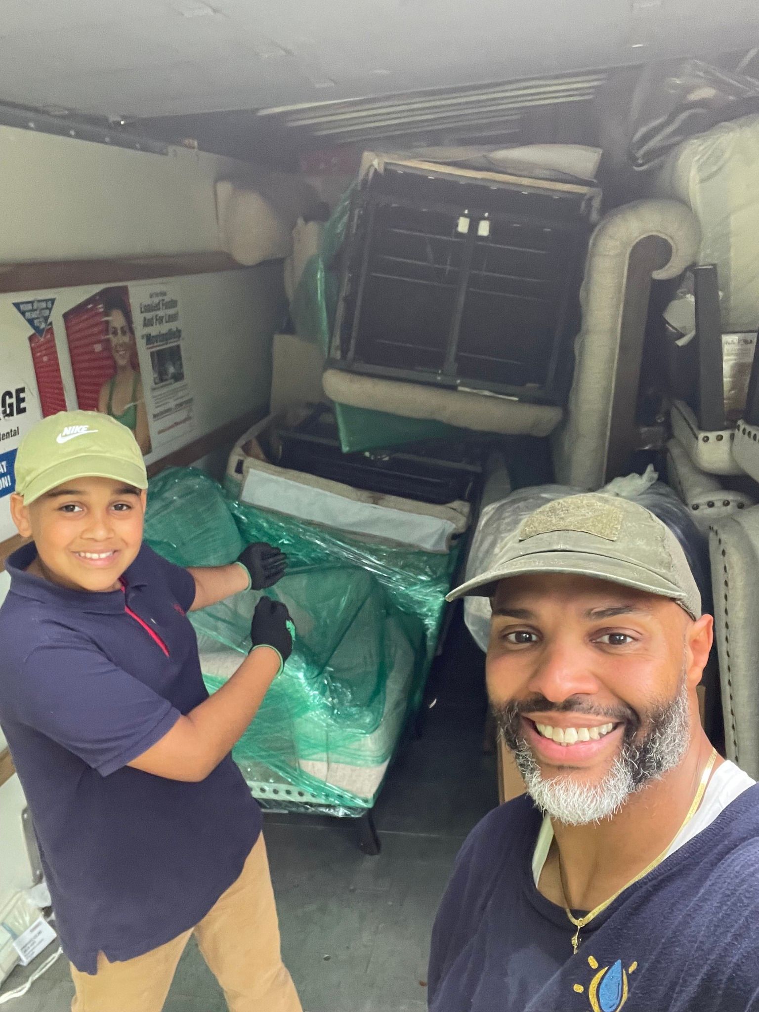 Two people smiling in a moving truck, loading furniture wrapped in green plastic.