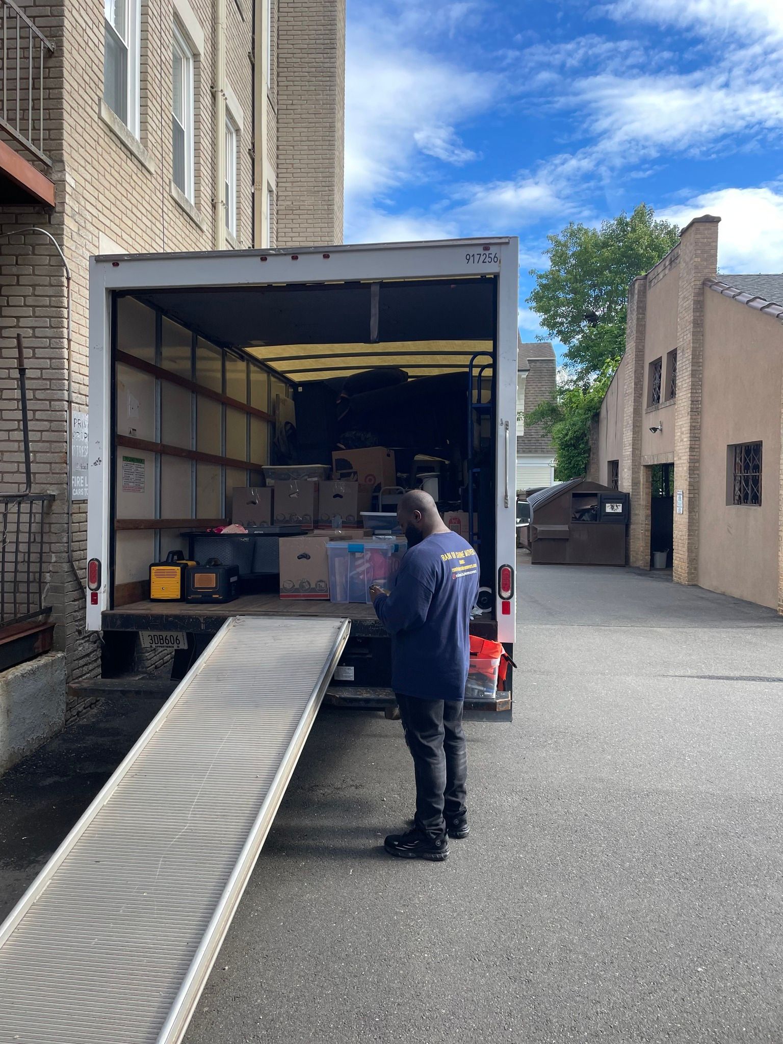 Man loading boxes into a moving truck, ramp extended.  Exterior shot, buildings and blue sky.