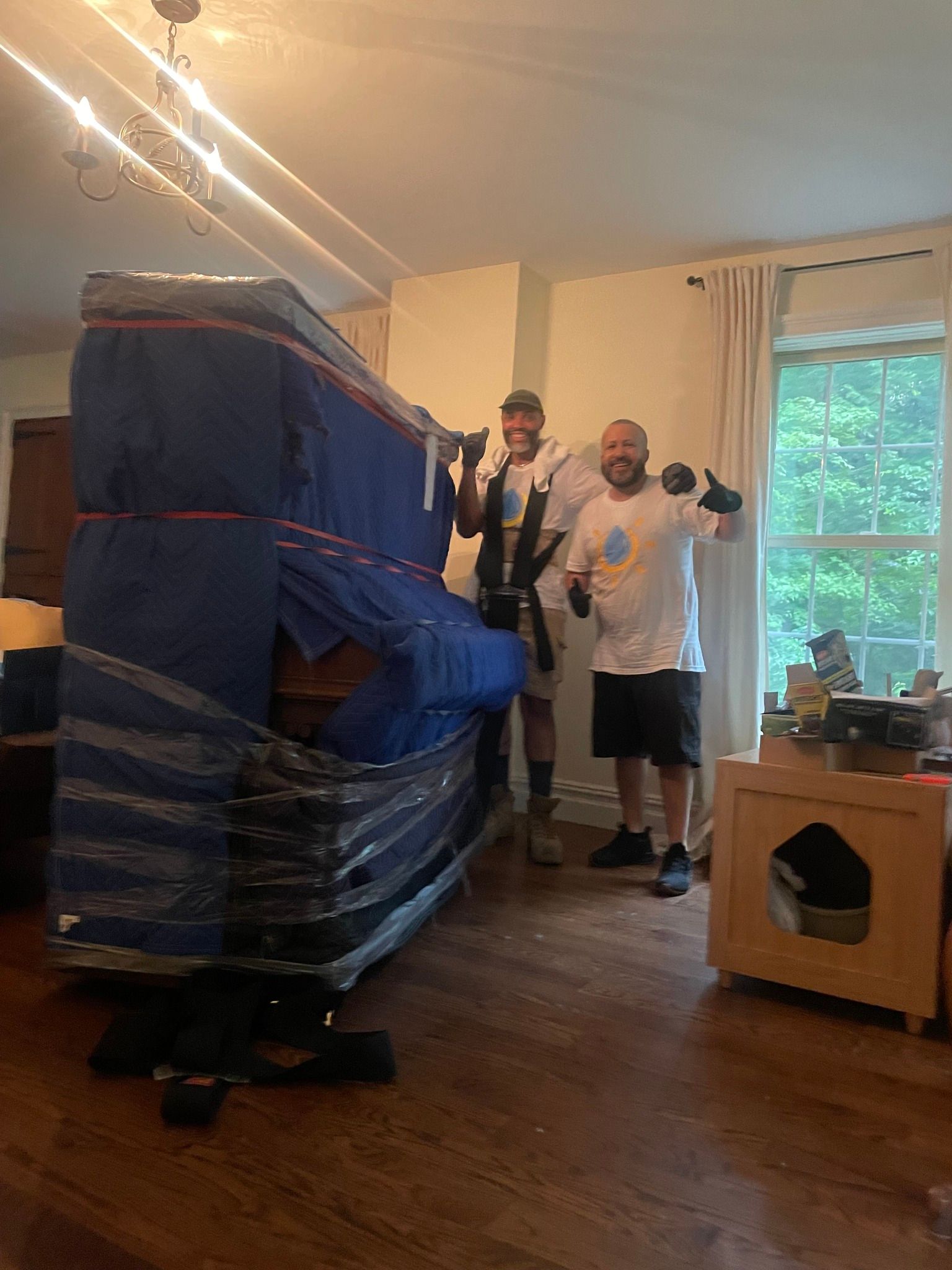 Two movers, smiling, next to a wrapped upright piano in a room with wood floors, window, and chandelier.
