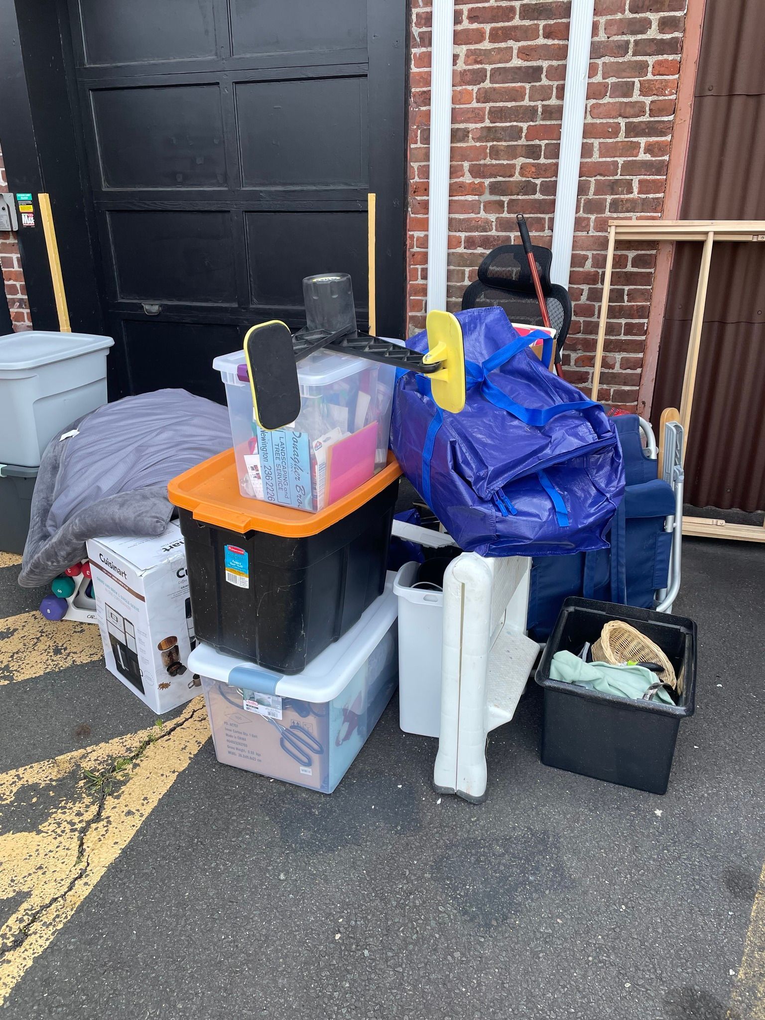 Outdoor shot of assorted storage bins, bags, and items piled against a brick building and black garage door.