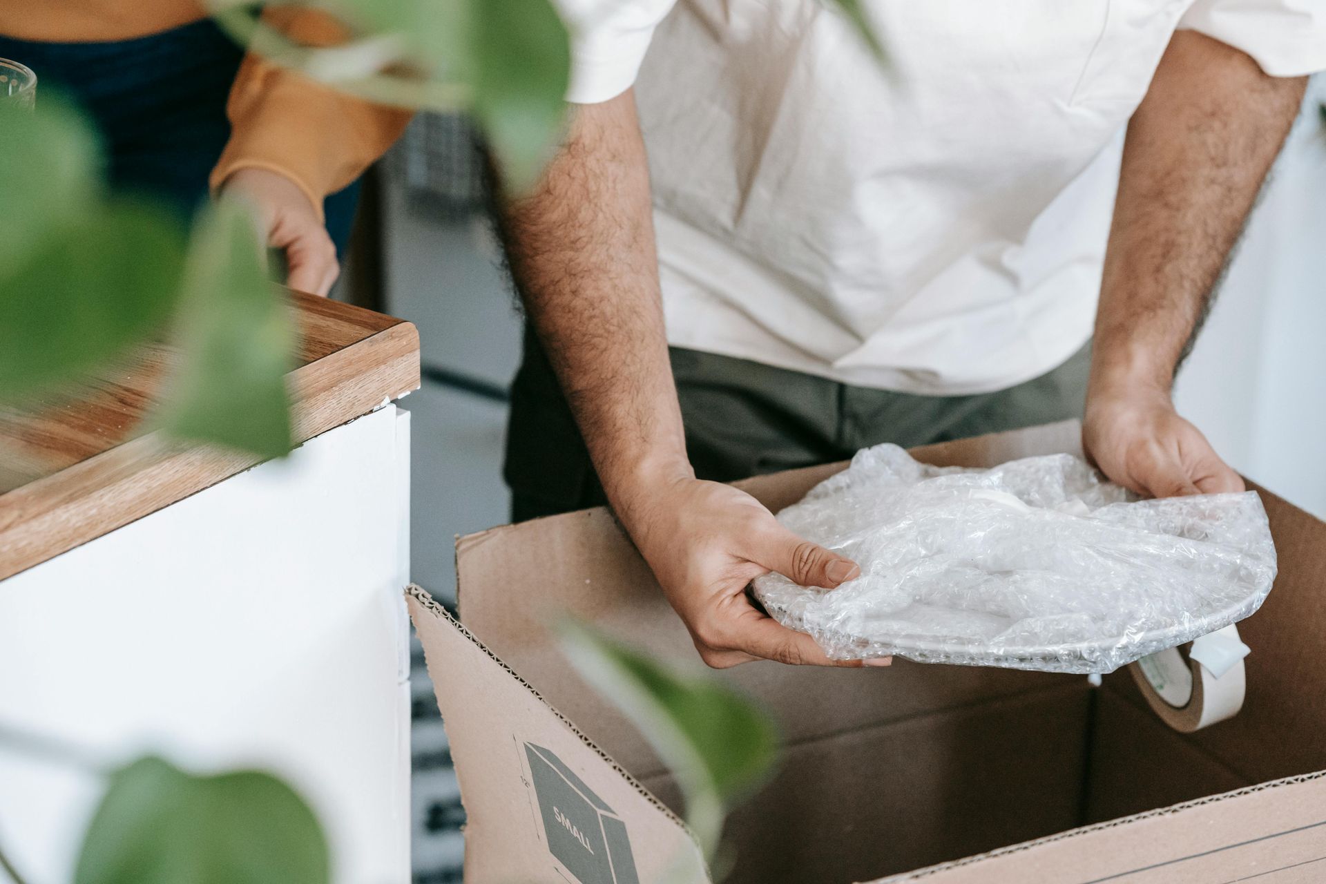Person packing a box, wrapping a dish with bubble wrap in a well-lit room.