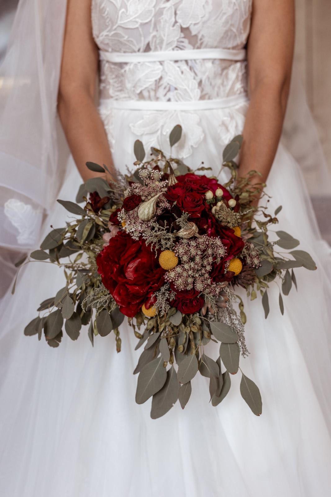 Eine Braut in einem weißen Kleid hält einen Strauß roter und gelber Blumen in der Hand.