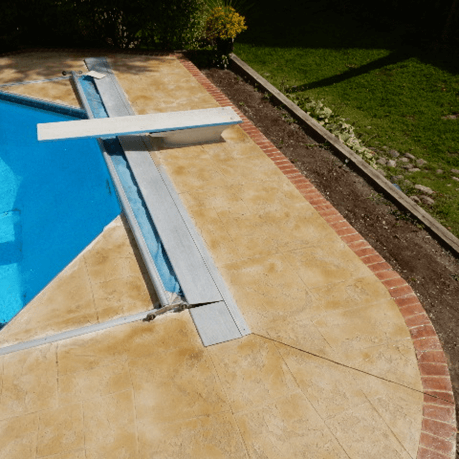 A diving board sits next to a swimming pool with stamped concrete and a decorative brick border around it