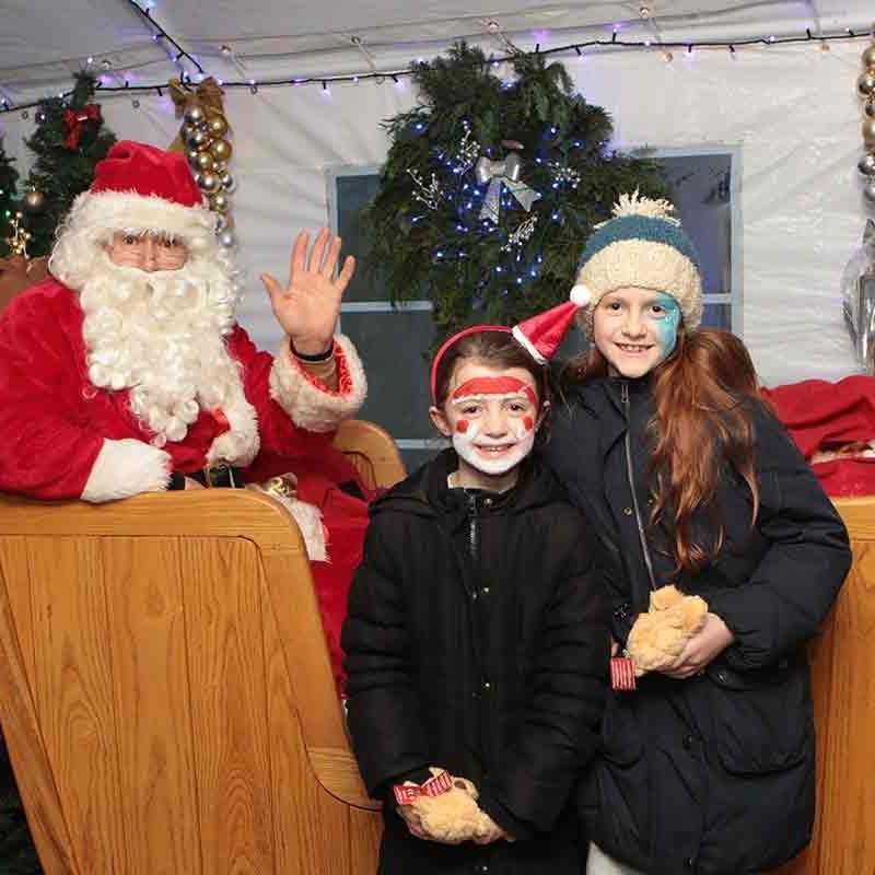 A festive scene inside Santa's grotto, with colourful decorations, twinkling lights, and Santa Claus seated in his chair surrounded by eager children.