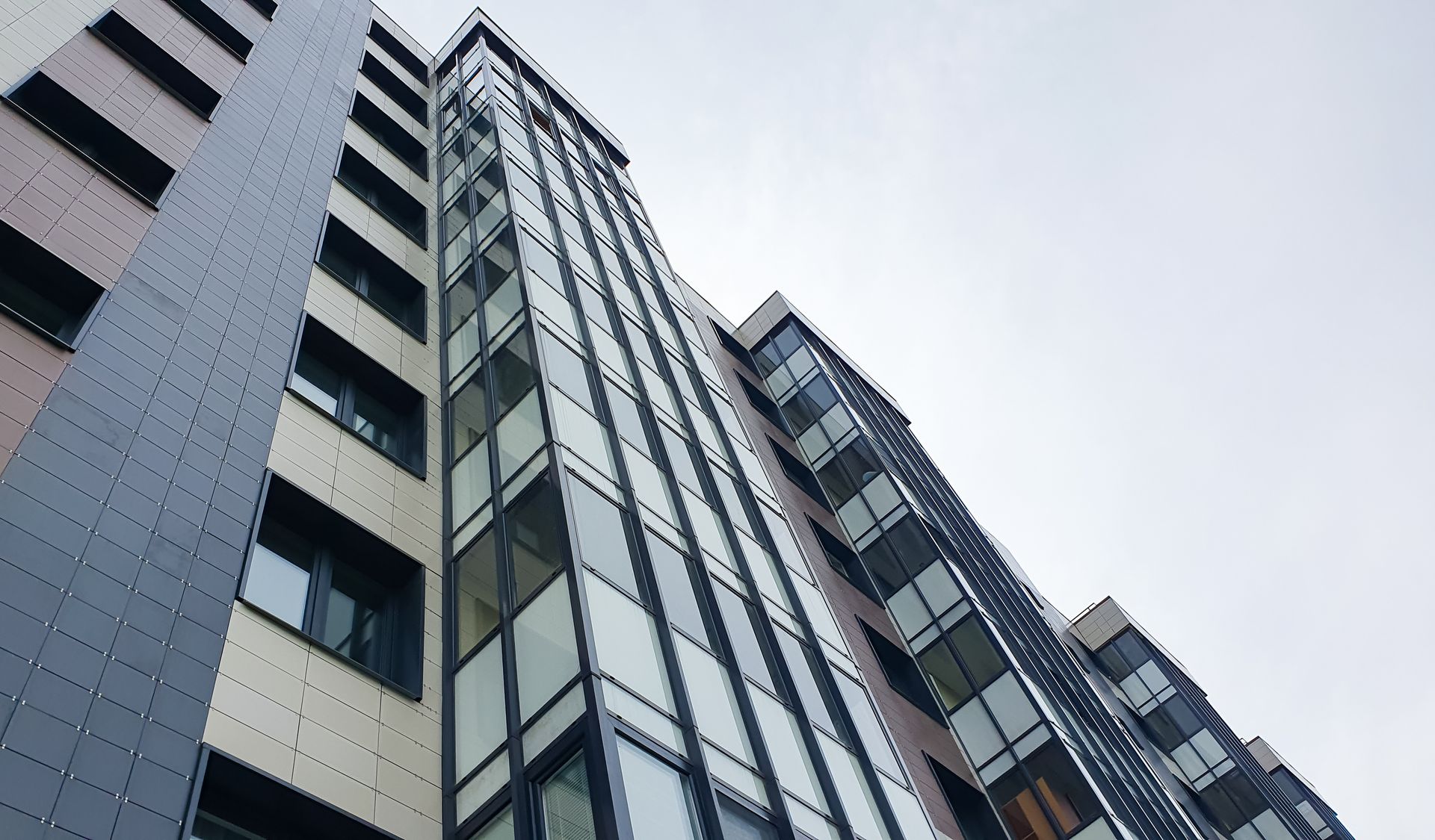 Tall modern building with dark and light panels, glass windows, angled upwards against cloudy sky.