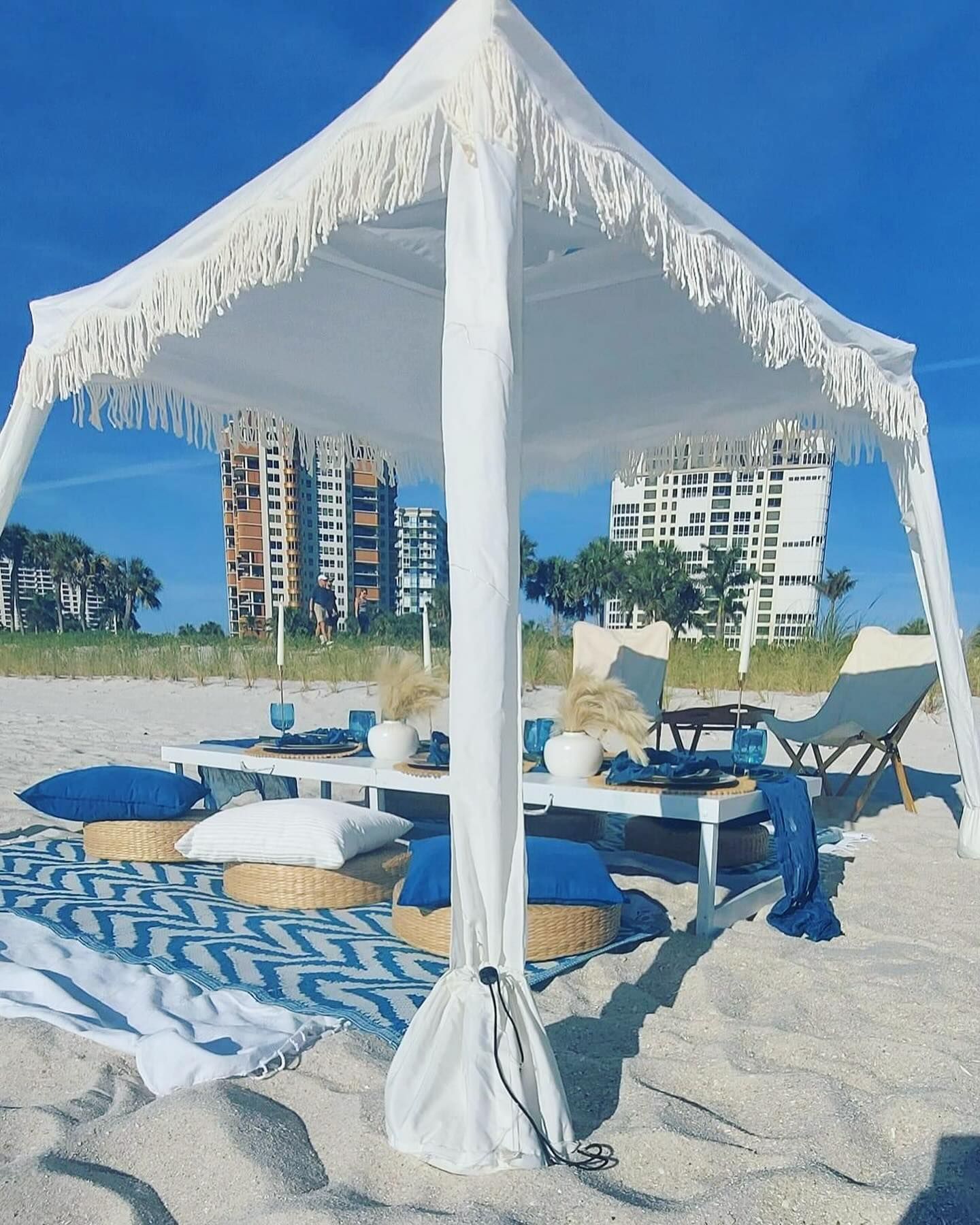 White canopy on a beach with picnic setup: table, pillows, blue decor. Buildings in the background.