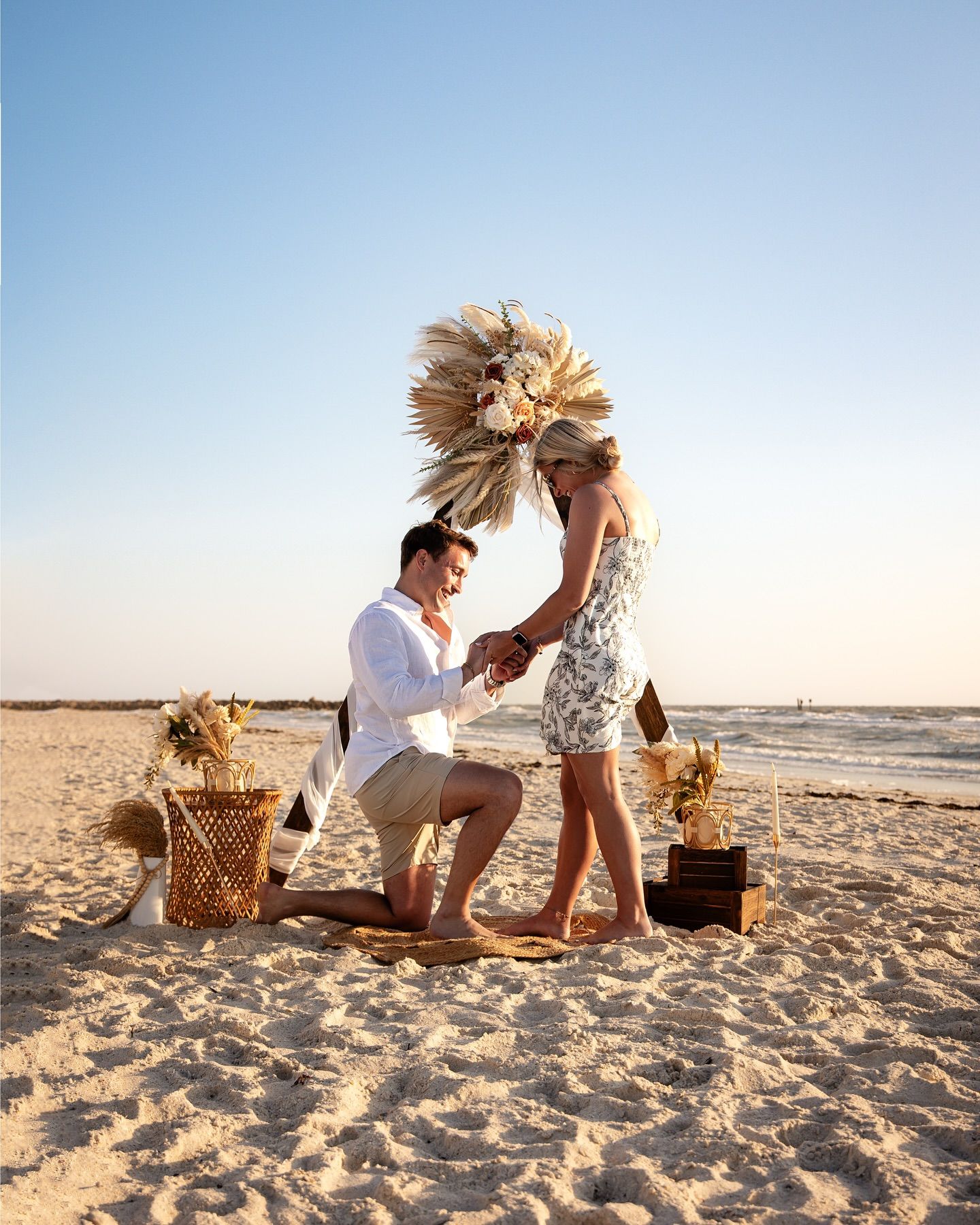 Man kneeling, proposing to woman on beach, under an arch decorated with flowers.