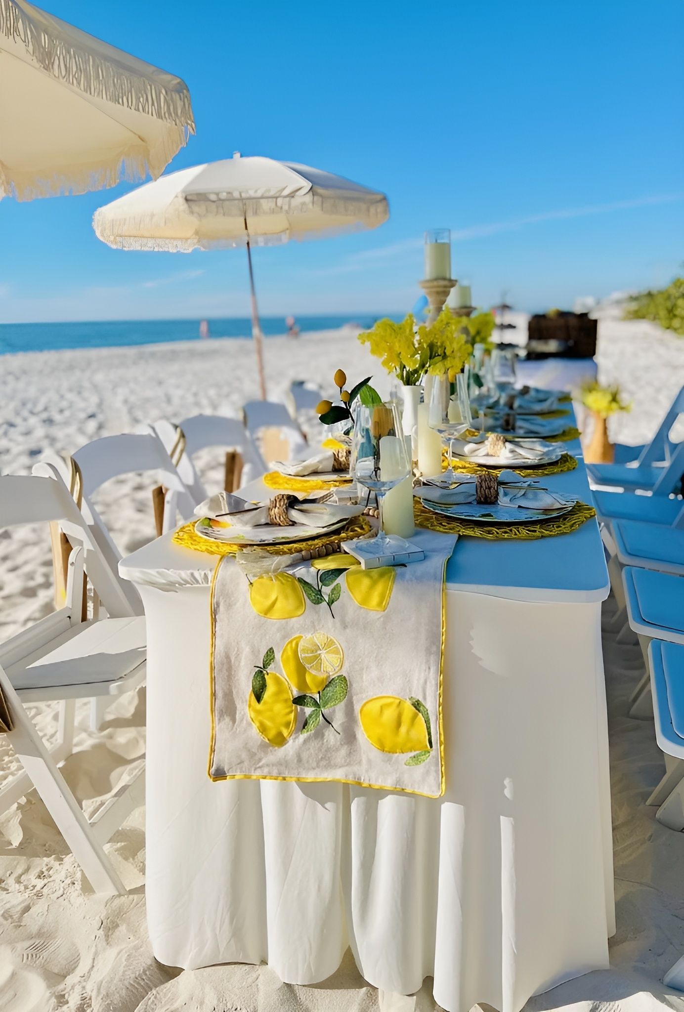 Beach table setting with lemons and yellow flowers under an umbrella.
