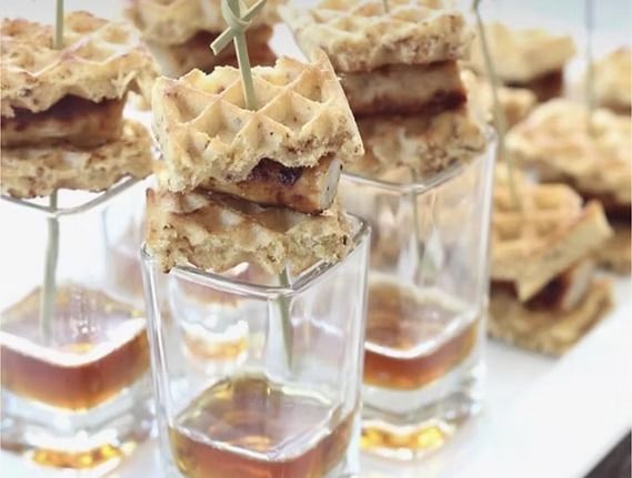 Mini waffle sandwiches skewered on sticks, served with syrup in small glasses.