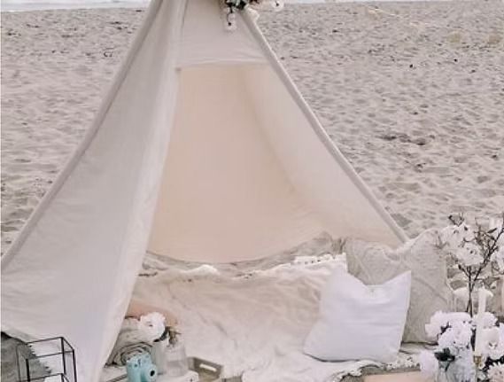 White teepee tent on a sandy beach, decorated with white pillows and flowers.