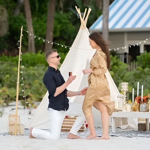 Man kneeling proposing to woman on beach, near teepee and table.