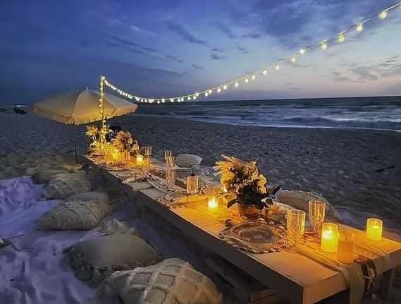 Beach picnic setup at dusk: long table, pillows, candles, string lights, and ocean.