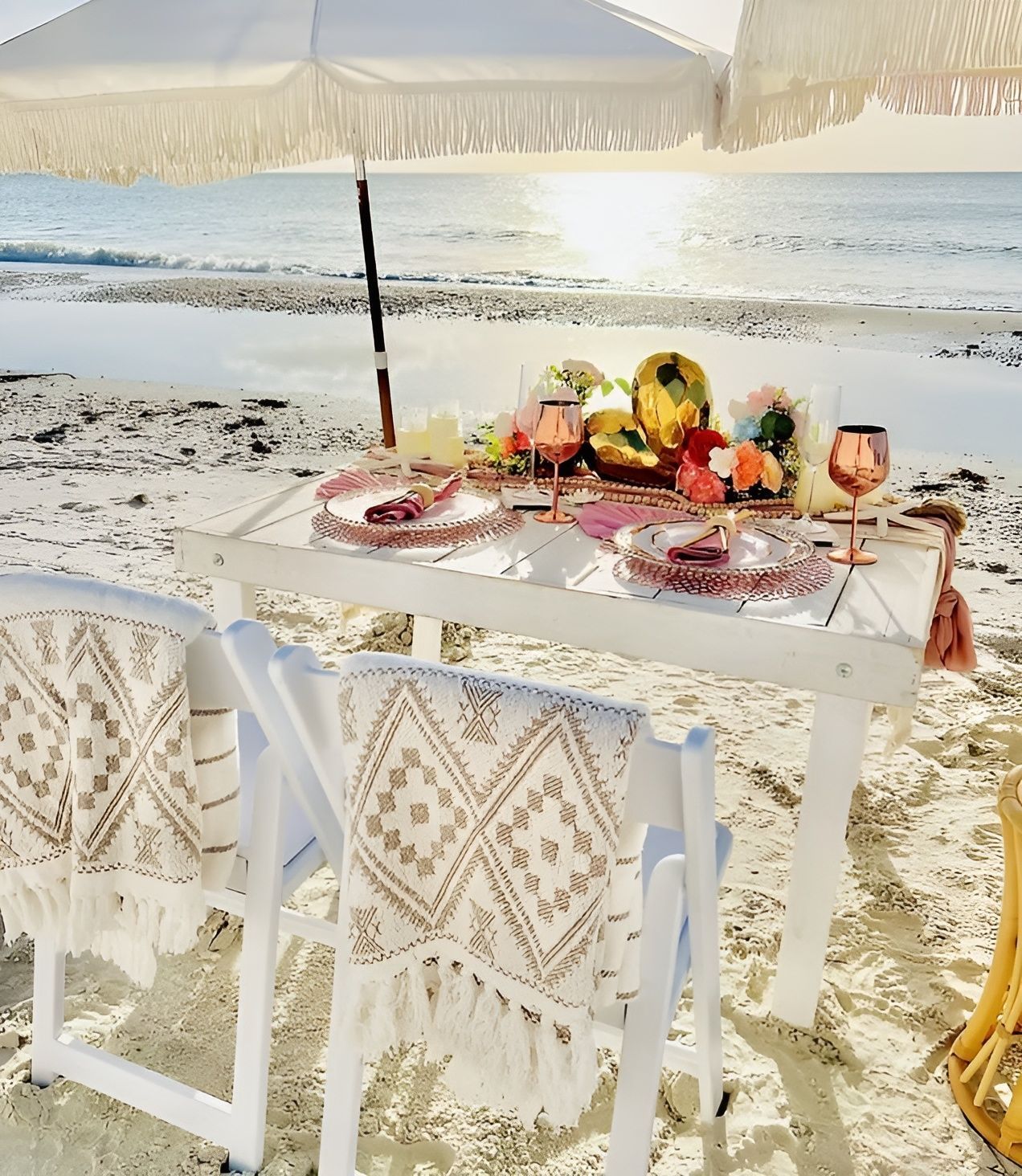 Beach picnic setup with white table, chairs, parasol, rose gold accents, and ocean backdrop.