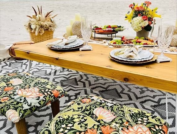 Picnic setup on a beach: wooden table with food and flowers, floral seating, patterned rug.