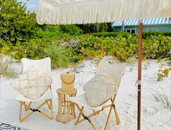 Beach scene: Two chairs with pillows, small table, umbrella, on sand.
