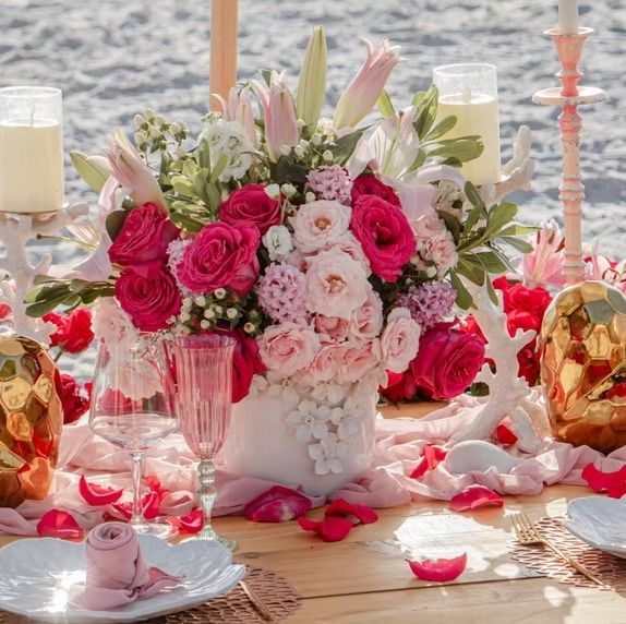 Romantic beach picnic with pink and red flowers, candles, and glassware on a wooden table.