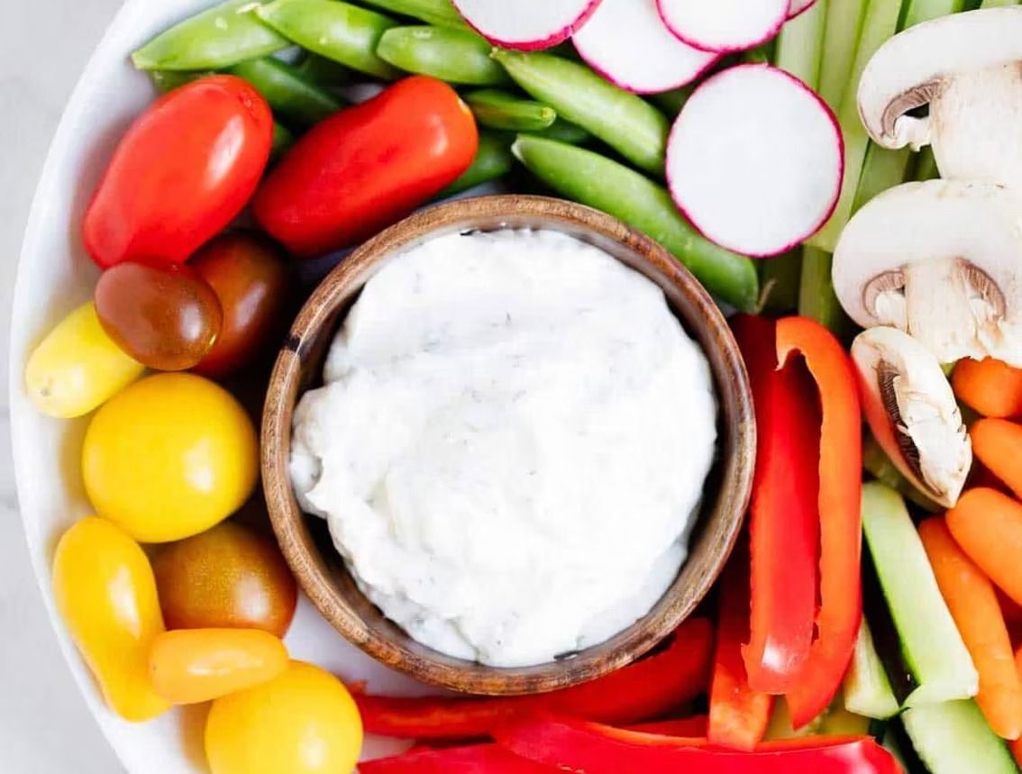 Vegetable platter with assorted colorful vegetables arranged around a wooden bowl of white dip.