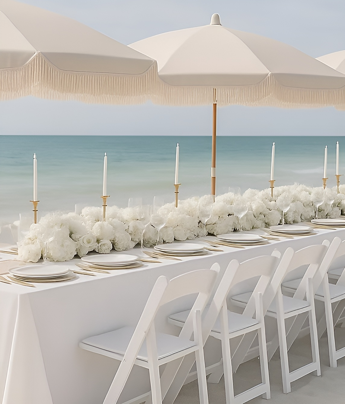 Beach table setting with white flowers, candles, umbrellas, and folding chairs. Ocean in background.