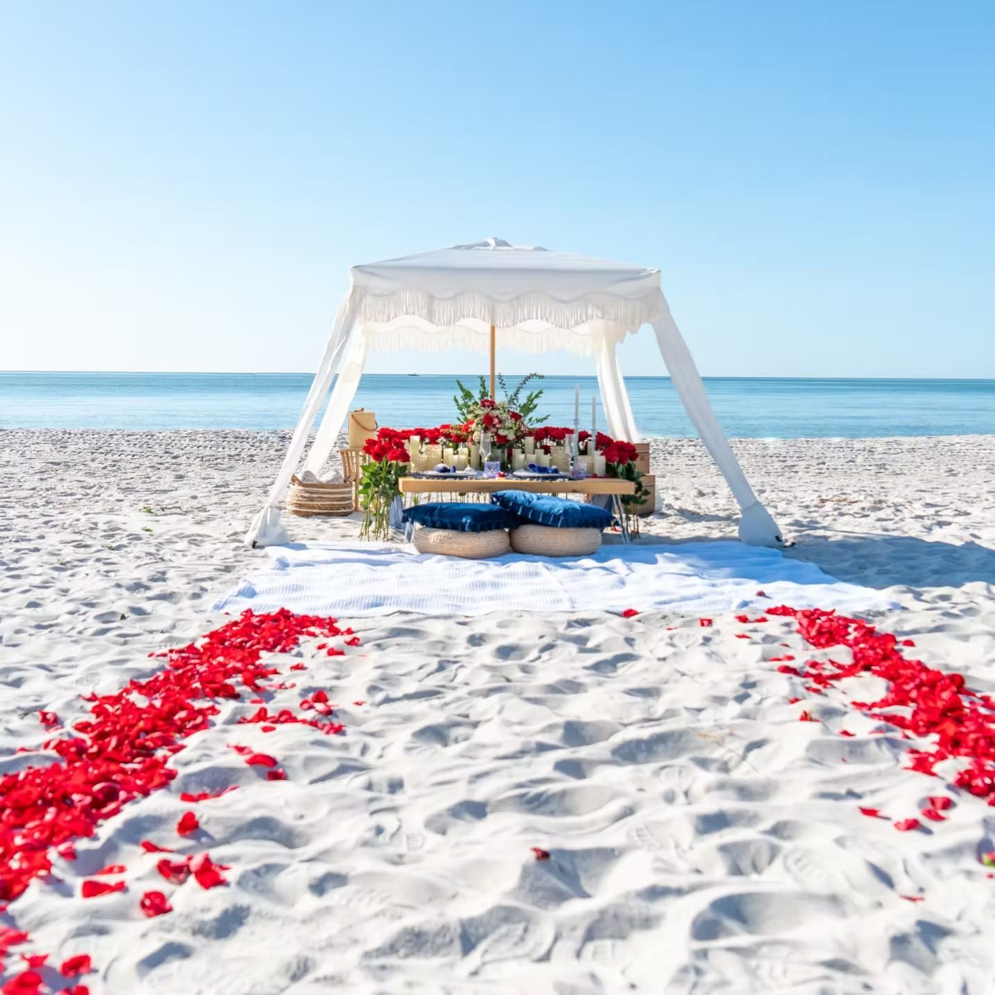 Beach setup with red rose petals, a table under a white canopy, and ocean view.