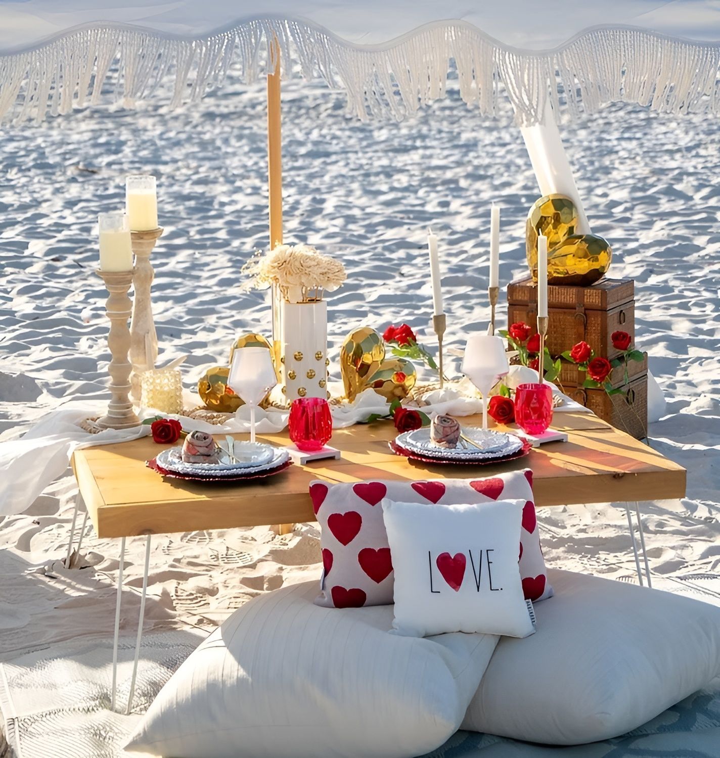 Beach picnic setup with table, pillows, candles, and decorative hearts.