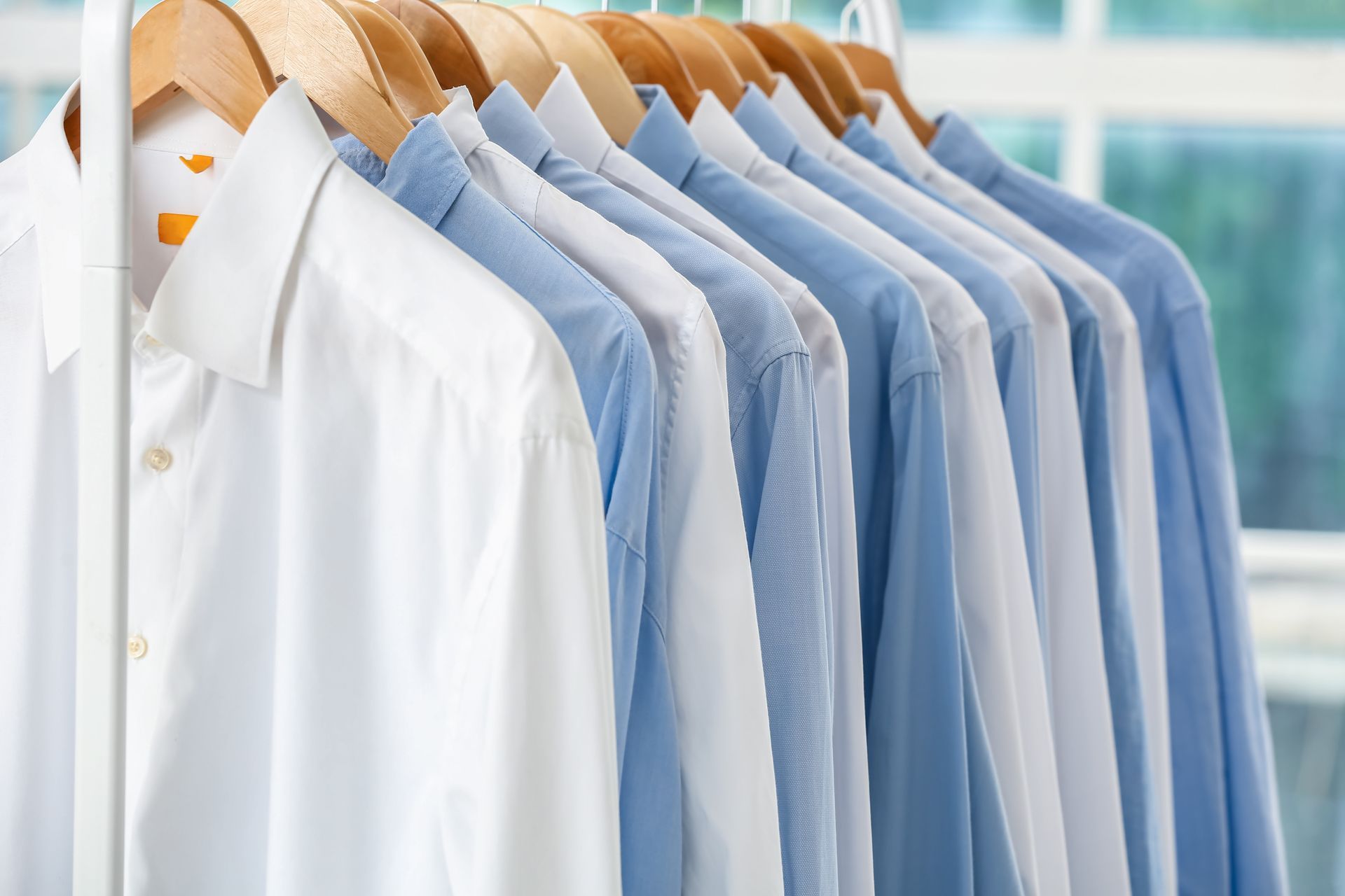 Row of button-down shirts on wooden hangers, varying shades of blue and white, hanging on a metal ra Row of button-down shirts on wooden hangers, varying shades of blue and white, hanging on a metal ra