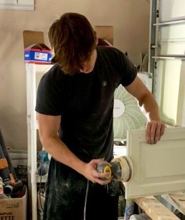 A man in a black shirt is sanding a white cabinet door.