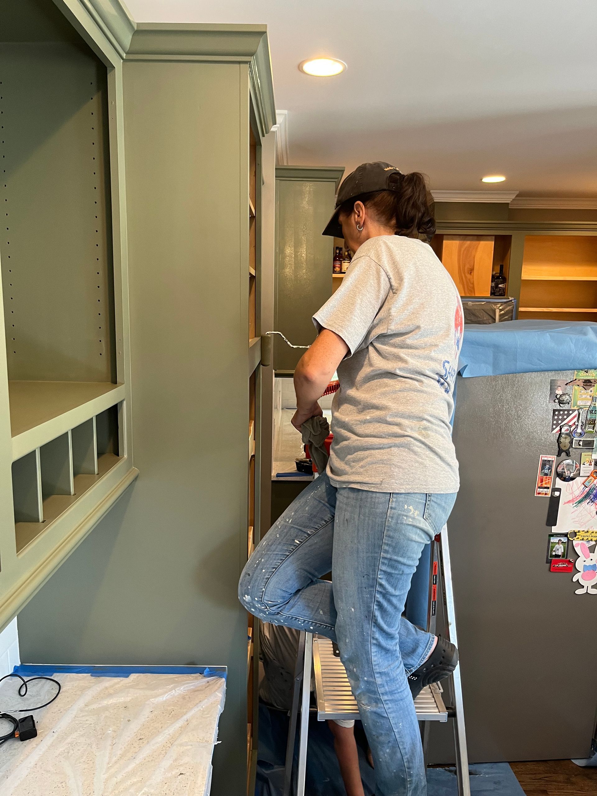 A woman is standing on a ladder working on a cabinet