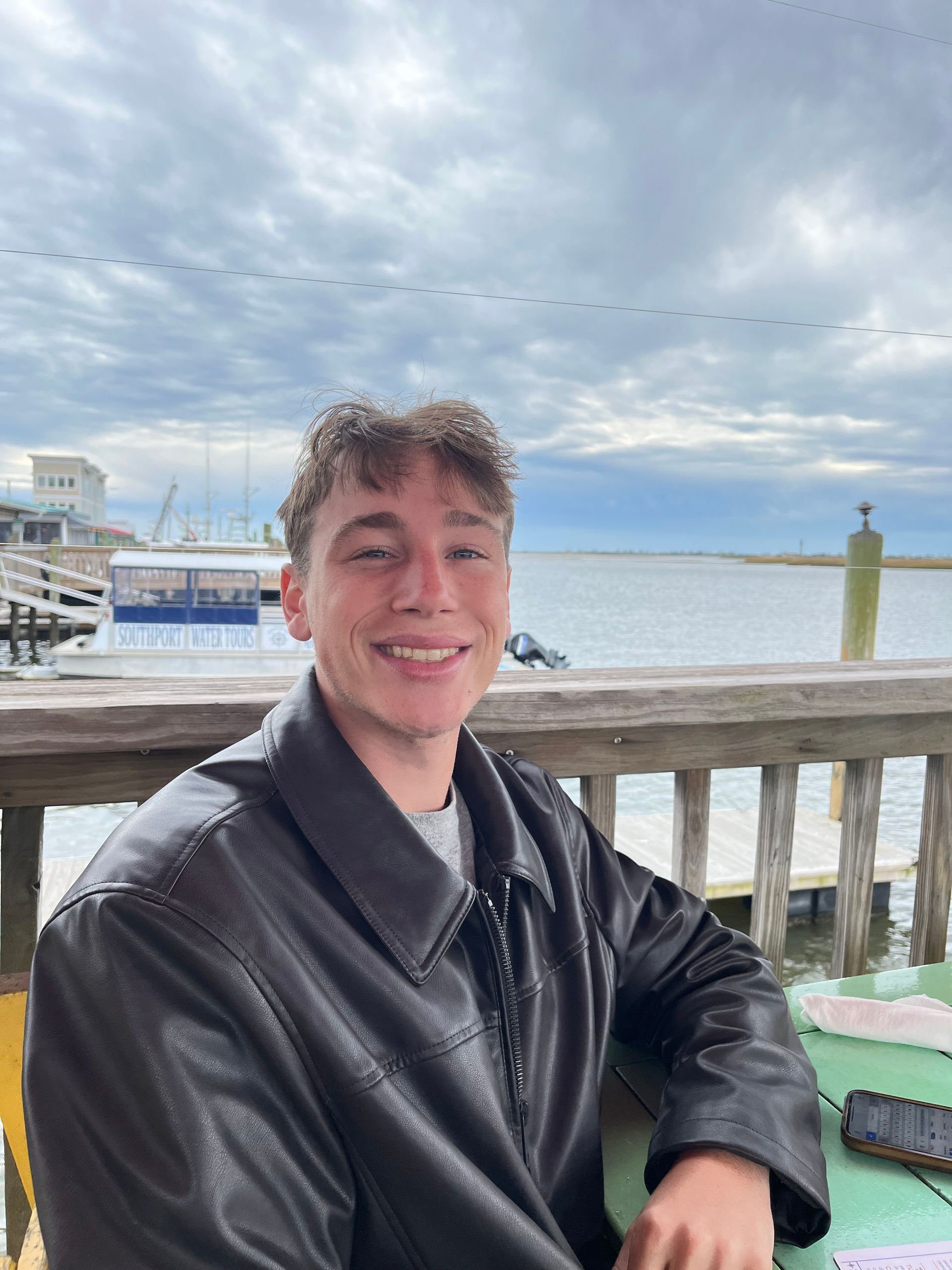 A young man in a leather jacket is sitting at a table in front of the ocean.