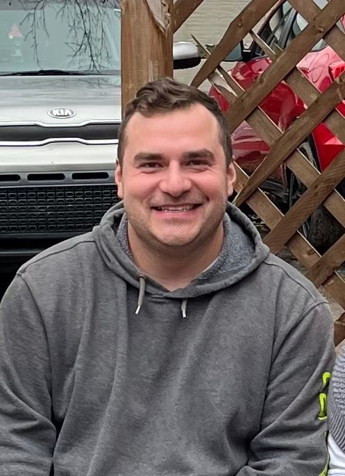 A man in a gray hoodie is smiling in front of a car.