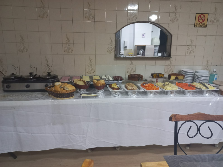 Mesa de bufê com diversos alimentos: bolos, pães e outros pratos, com pratos empilhados à direita.